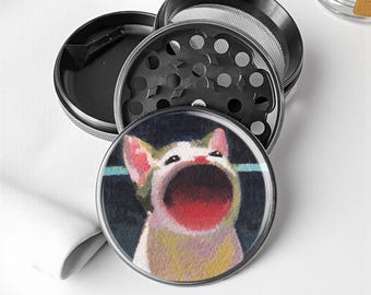 May include: A black and silver herb grinder with a cat illustration on the lid. The cat has a wide open mouth and is painted in shades of white, yellow, and red. The grinder is disassembled, showing the different parts.
