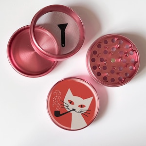 Op de afbeelding: Een gedemonteerde roze metalen kruidenmolen met een gaasscherm, een kleine zwarte schraper en een deksel met een kat die een pijp rookt. De kat is wit met rode ogen op een rode achtergrond.