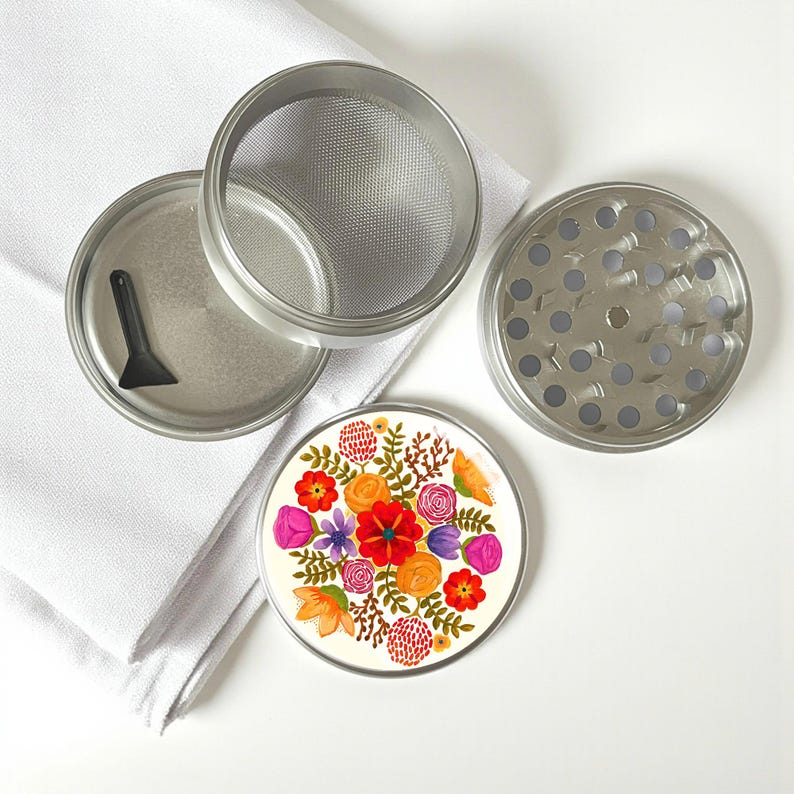 May include: A disassembled, stainless steel herb grinder with a floral design. The grinder includes a lid, a mesh screen, a grinding plate, and a base with a colorful floral pattern featuring red, orange, and purple flowers. A small black scraper is included.