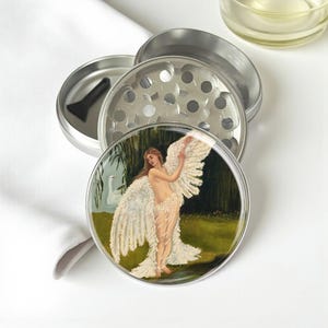 May include: A silver metal herb grinder with a detailed illustration of a woman with angel wings. The artwork features a woman in a flowing gown with white wings, standing near a body of water with a swan.