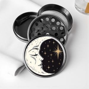 May include: A black and silver four-piece herb grinder. The top piece features a design of a crescent moon with a face, and a starburst and stars on a black background. The grinder is made of metal.