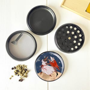 May include: A four-piece black herb grinder with a whimsical illustration of dancing figures on the bottom piece. The grinder includes a screen, a small scraper, and a top and bottom chamber. A small pile of herbs is visible.