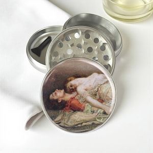 May include: A silver metal herb grinder with a detailed painting on the lid. The painting depicts two figures in a classical style. The grinder is partially disassembled, revealing the grinding components and a small black scraper.