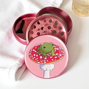 Peut inclure: Un grinder à herbes rose avec un motif fantaisiste. Le dessus du grinder présente une grenouille de dessin animé assise sur un champignon rouge à pois blancs. Le grinder est démonté, révélant les composants et un petit grattoir noir.