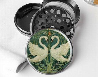 May include: A black, four-piece herb grinder with a decorative top featuring two white swans facing each other against a green and gold background. The grinder is made of metal and has a mesh screen.