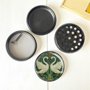 May include: A four-part black herb grinder featuring a swan design. The grinder comprises a top, a grinding chamber, a mesh screen with a small scraper, and a base piece with a swan illustration. The swan illustration shows two white swans with green and gold detailing.