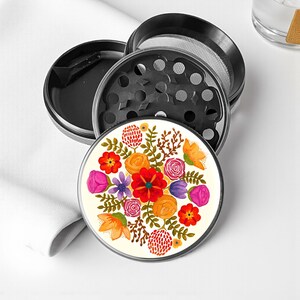 May include: A multi-piece, black and silver herb grinder with a floral design. The top piece features a cream-colored surface with a vibrant watercolor floral pattern in red, orange, purple, and yellow. The grinder is designed for herbs and spices.