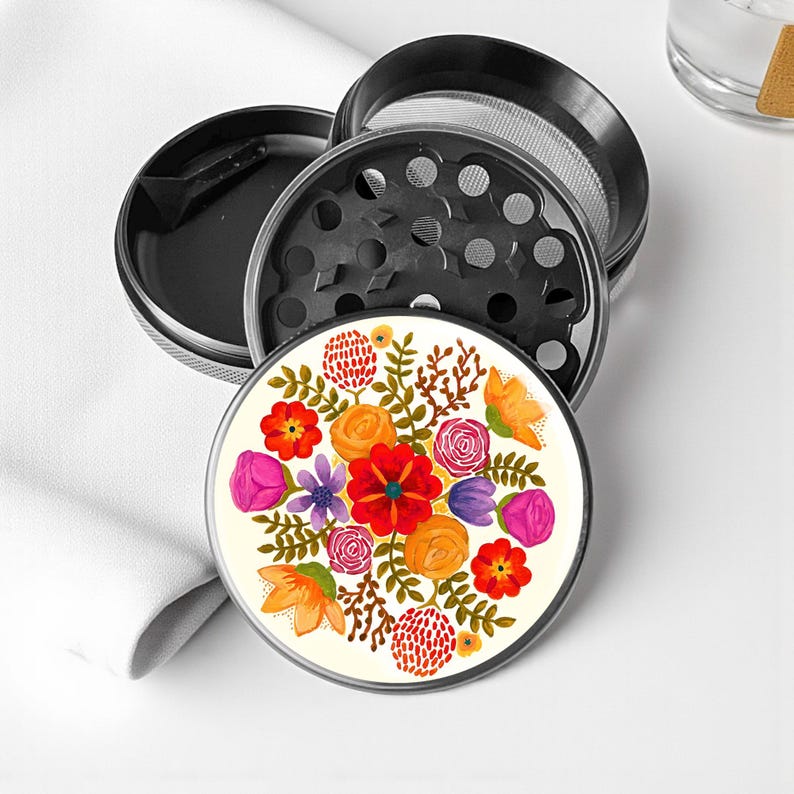 May include: A multi-piece, black and silver herb grinder with a floral design. The top piece features a cream-colored surface with a vibrant watercolor floral pattern in red, orange, purple, and yellow. The grinder is designed for herbs and spices.