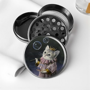 May include: A four-piece, black and silver herb grinder. The bottom piece features a whimsical illustration of a cat wearing a crown and a pink outfit, blowing bubbles. The grinder is designed for grinding herbs and spices.