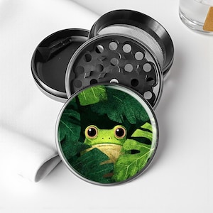 May include: A disassembled, four-piece herb grinder. The top piece features a frog peeking through green leaves. The grinder is black and silver, with a mesh screen and a compartment for ground herbs.