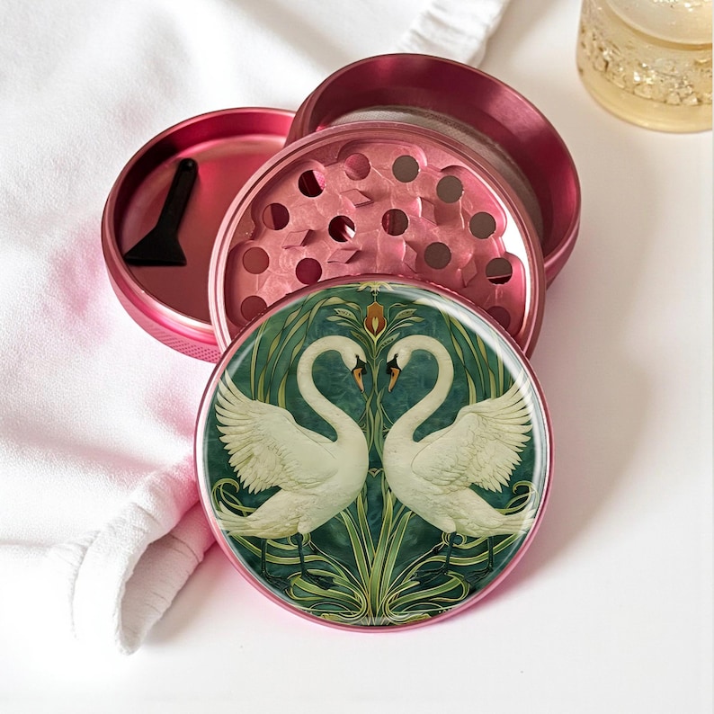 May include: A pink metal grinder with a decorative lid featuring two white swans facing each other against a green and teal background. The grinder is open, revealing the grinding components and a small black scraper.