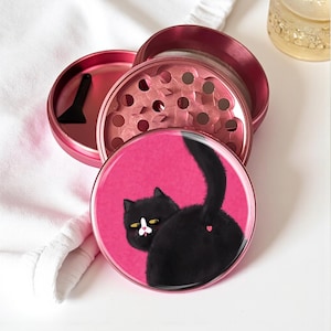 May include: A disassembled pink metal grinder with a black plastic scraper. The grinder's top features a pink graphic of a black cat with a pink heart on its rear end, set against a pink background.