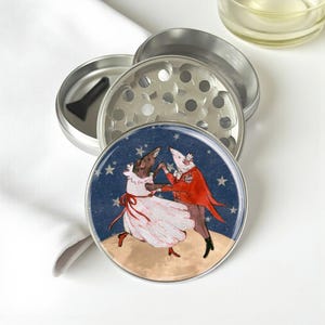 May include: A silver metal herb grinder with a whimsical illustration of two mice dancing on the lid. The mice are dressed in formal attire against a starry blue background. Includes a black scraper tool.