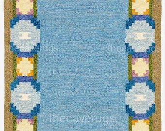 Op de afbeelding: Blauw rechthoekig tapijt met een patroonrand. De rand heeft geometrische vormen in blauw, geel en paars tegen een bruine achtergrond. Het tapijt heeft gefranjerde randen en de tekst "thecaverugs".