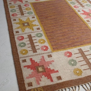 Puede incluir: Alfombra rectangular tejida a mano con un centro marrón y un borde crema. El borde presenta patrones geométricos en rojo, amarillo, verde y marrón. La alfombra tiene un borde con flecos y el texto "@thecaverugs".