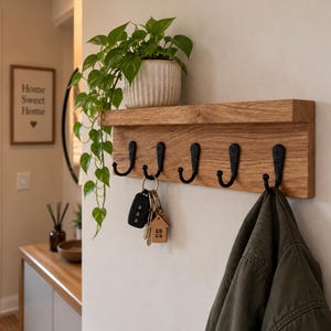 May include: A light brown wooden wall-mounted shelf featuring five black metal hooks. A green potted plant sits on the top ledge. A set of keys with a small house-shaped keychain hangs from one hook, and a dark green jacket hangs from another hook.