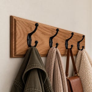 Könnte beinhalten: Eine Wandgarderobe mit einem rechteckigen Eichenholzbrett und vier mattschwarzen Doppelhaken aus Metall. An den Haken hängen eine olivgrüne Jacke, ein beiger Strickschal, eine braune Ledertasche und eine cremefarbene, strukturierte Fleecejacke.