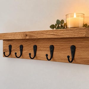 May include: A wall-mounted wooden shelf featuring a flat top surface and a vertical back panel. Five black metal hooks are attached to the front of the panel. A glass candle holder with a lit white candle and green leaves sits on the shelf.