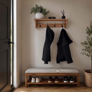 May include: A wooden wall-mounted coat rack with four black metal hooks and a top shelf holding a white ribbed vase with greenery, sunglasses, and a reed diffuser. Below, a wooden bench with a light grey upholstered seat and a slatted shoe rack.