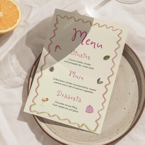 Może przedstawiać: Karta menu z jasnobeżowym tłem i różowo-zieloną falistą obwódką. Menu zawiera sekcje dla przystawki, dania głównego i deserów z opisami potraw. Widoczna jest również pomarańcza przecięta na pół i talerz.