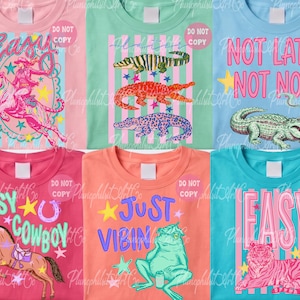 May include: Collection of colorful t-shirts with graphic designs. Designs include a woman riding a tiger, alligators, a frog, a horse, and a tiger. Each shirt has text like "EASY", "NOT LATER NOT NOW", and "JUST VIBIN".
