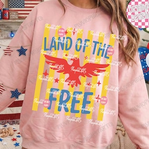 Puede incluir: Una sudadera rosa claro con un dise&ntilde;o patri&oacute;tico. El dise&ntilde;o presenta las palabras "LAND OF THE FREE" en azul, un &aacute;guila roja, rayas amarillas y estrellas rojas. La sudadera es de estilo c&oacute;modo e informal.