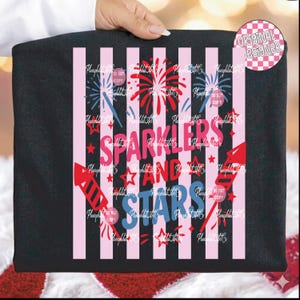 Puede incluir: Camiseta negra con un dise&ntilde;o patri&oacute;tico. Presenta rayas rosas y blancas, fuegos artificiales, estrellas y las palabras "Sparklers and Stars" en rosa y azul. Un logotipo circular de "Dise&ntilde;ador original" est&aacute; en la esquina superior derecha.