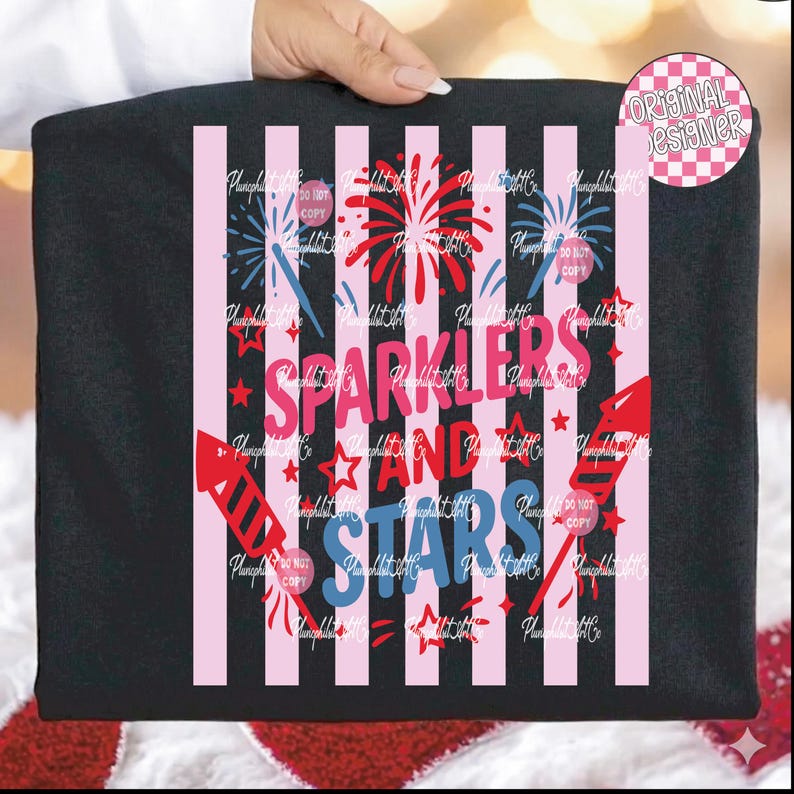 Puede incluir: Camiseta negra con un dise&ntilde;o patri&oacute;tico. Presenta rayas rosas y blancas, fuegos artificiales, estrellas y las palabras "Sparklers and Stars" en rosa y azul. Un logotipo circular de "Dise&ntilde;ador original" est&aacute; en la esquina superior derecha.