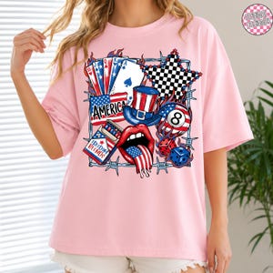 May include: Pink t-shirt with a patriotic graphic featuring playing cards, a star, a top hat, an eight ball, dice, and a mouth with a tongue shaped like the American flag. The word "AMERICA" is also displayed.