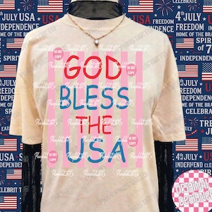 Puede incluir: Camiseta color crema con el texto "GOD BLESS THE USA" en rojo y azul, con rayas rosas y azules. La camiseta tiene un dise&ntilde;o patri&oacute;tico, ideal para celebrar el D&iacute;a de la Independencia.