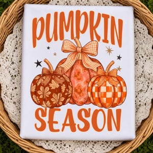 May include: A white t-shirt featuring the orange text PUMPKIN SEASON. The design includes three pumpkins with orange gingham bows. The left pumpkin has a brown leaf pattern, the center is solid orange, and the right has an orange checkerboard pattern.