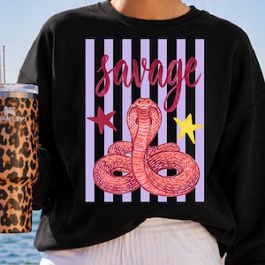 May include: Black sweatshirt featuring a pink cobra illustration with the word "Savage" in pink script. The design includes a purple striped background, a red star, and a yellow star.