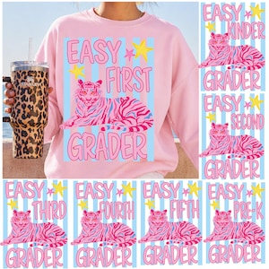 Puede incluir: Sudadera rosa con un diseño de tigre rosa y azul y la frase "Easy First Grader". En la imagen se sostiene un vaso con estampado de leopardo. Los diseños adicionales incluyen "Easy Kinder Grader", "Easy Second Grader", "Easy Third Grader", "Easy Fourth Grader", "Easy Fifth Grader" y "Easy Pre-K Grader".