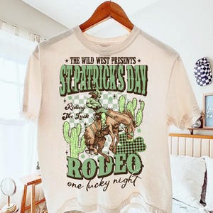 Puede incluir: Camiseta color crema con un diseño de rodeo del Día de San Patricio. El gráfico presenta un vaquero montando un caballo salvaje, cactus y el texto "St. Patrick's Day Rodeo, one lucky night". La camiseta está colgada en una percha de madera.