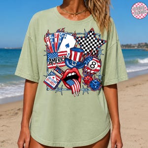 May include: Sage green t-shirt featuring a patriotic graphic with playing cards, a top hat, an eight ball, dice, and a star. The word "AMERICA" is displayed prominently. The design incorporates red, white, and blue colors, and the text "FREEDOM BLENDS" is also visible.