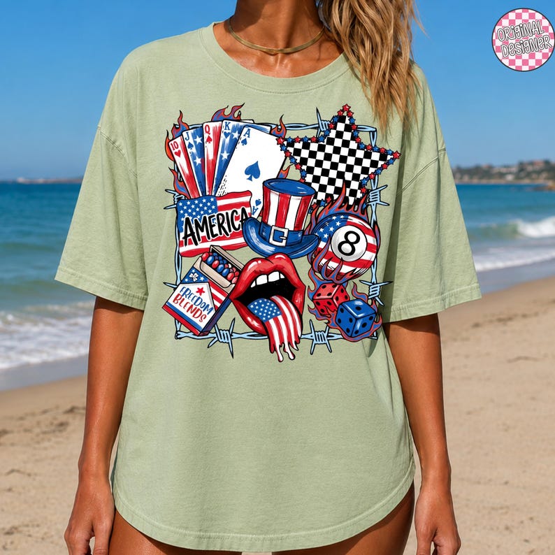 May include: Sage green t-shirt featuring a patriotic graphic with playing cards, a top hat, an eight ball, dice, and a star. The word "AMERICA" is displayed prominently. The design incorporates red, white, and blue colors, and the text "FREEDOM BLENDS" is also visible.