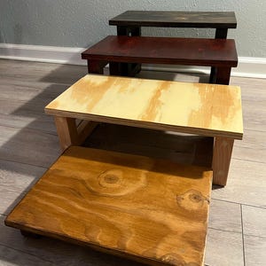 May include: Four wooden step stools of varying heights and colors are stacked. The top stool is dark brown, the second is a deep red, the third is light yellow, and the bottom stool is a natural wood color. The stools are made of wood and have a simple design.