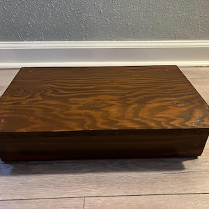 May include: A square, wooden footstool with a dark brown finish. The wood grain is visible across the top surface. The stool has a simple, rectangular design and sits directly on the floor.