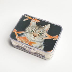 May include: A clear plastic pill box with a hinged lid. The lid features a grumpy-looking grey tabby cat with orange goldfish swimming around its head. The background is black.