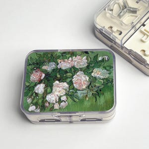 May include: A rectangular, clear plastic pill organizer with a floral design on the lid. The design features a painting of white and pink roses with green foliage against a green background. Another organizer is partially visible.