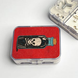May include: A clear plastic container with a red felt insert. The insert features a cartoon illustration of a poison bottle. The bottle is black with a skull and crossbones design, a blue neck, and a cork stopper.
