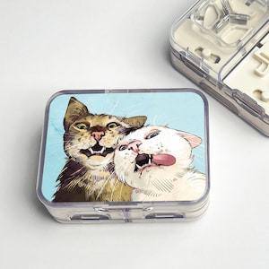 Pode incluir: Um organizador de comprimidos de plástico transparente com uma ilustração de desenho animado de dois gatos na tampa. Um gato é castanho e o outro branco, ambos com expressões exageradas sobre um fundo azul claro.