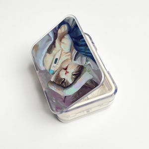 May include: A clear, hinged pill case with a painted image of a cat lying down with a thermometer. The cat is resting on a bed of blankets and pillows. The case is open, revealing the interior compartments.