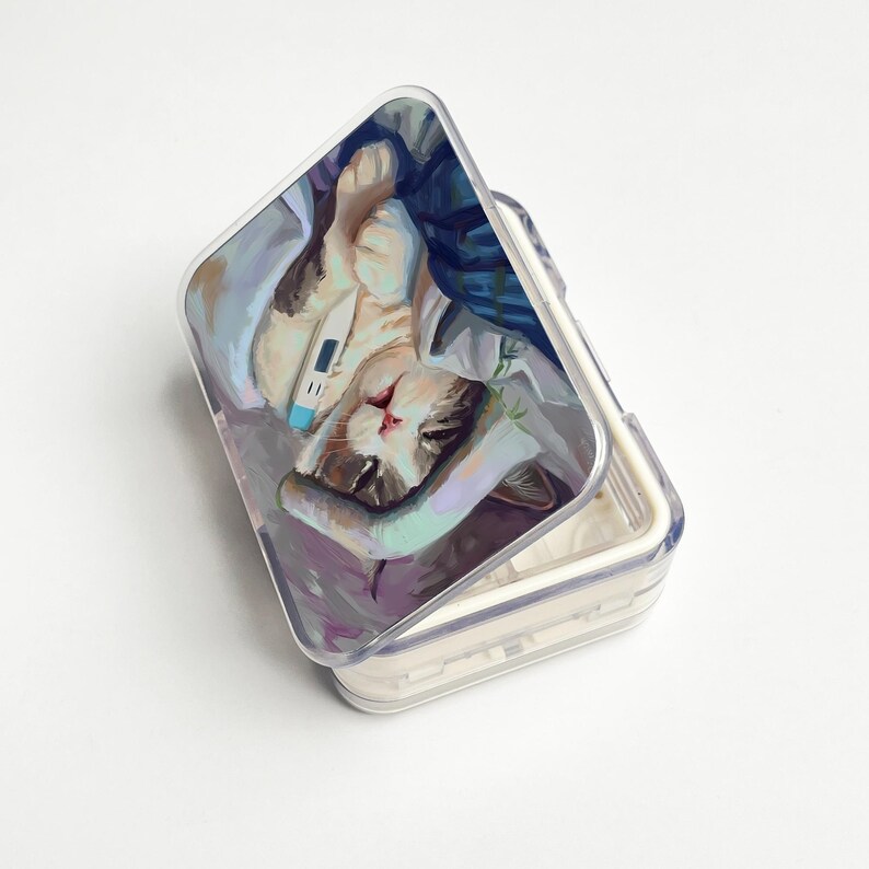 May include: A clear, hinged pill case with a painted image of a cat lying down with a thermometer. The cat is resting on a bed of blankets and pillows. The case is open, revealing the interior compartments.