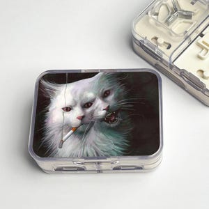 Pode incluir: Uma caixa de plástico transparente com um gráfico de um gato branco de duas faces, um lado fumando um cigarro, o outro rosnando. A caixa é retangular e tem uma tampa articulada. Outra caixa de plástico transparente é parcialmente visível.