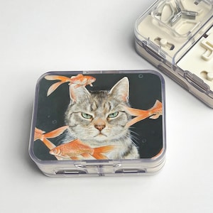 May include: A clear plastic pill case featuring a cat and goldfish design. The cat has a stern expression, surrounded by orange goldfish. The case is rectangular with a hinged lid and a white interior.