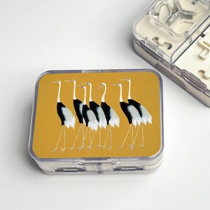 May include: A clear plastic container with a yellow lid featuring a design of six black and white cranes. The cranes are standing on long legs against a yellow background. The container is rectangular.