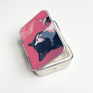 May include: A clear plastic pill case with a pink lid featuring a graphic of a cat with a cigarette. The cat is black, white, and grey, with a cigarette held by a hand above its head. Smoke rises from the cigarette.