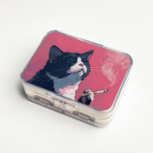 May include: A clear, rectangular pill case with a pink lid featuring a black and white cat smoking a cigarette. The cat is depicted with closed eyes and a relaxed expression, with smoke rising from the cigarette.