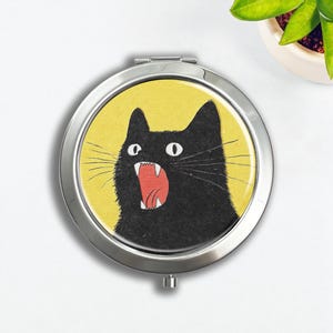 Op de afbeelding: Een ronde, zilveren compacte spiegel met een cartoonillustratie van een zwarte kat met open mond op een gele achtergrond. De kat heeft witte ogen en een rode tong. De compact is open en toont de spiegel.